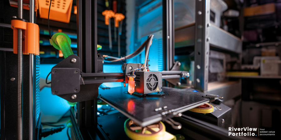 A close-up, dynamic shot of a 3D printer in operation, with its print head actively extruding material onto the build plate. The machine's internal mechanisms, including motors and belts, are visible, illuminated by a blue glow from within. The background is blurred, showing a workshop or lab environment with various tools and equipment, suggesting an innovative and creative space. In the bottom right corner, the "RiverView Portfolio | Your added value accountants" logo is present, indicating the image's relevance to research and development activities and potential R&D tax credit claims. Property Tax Advice Expat Tax Advice PTA ETA Riverview Portfolio