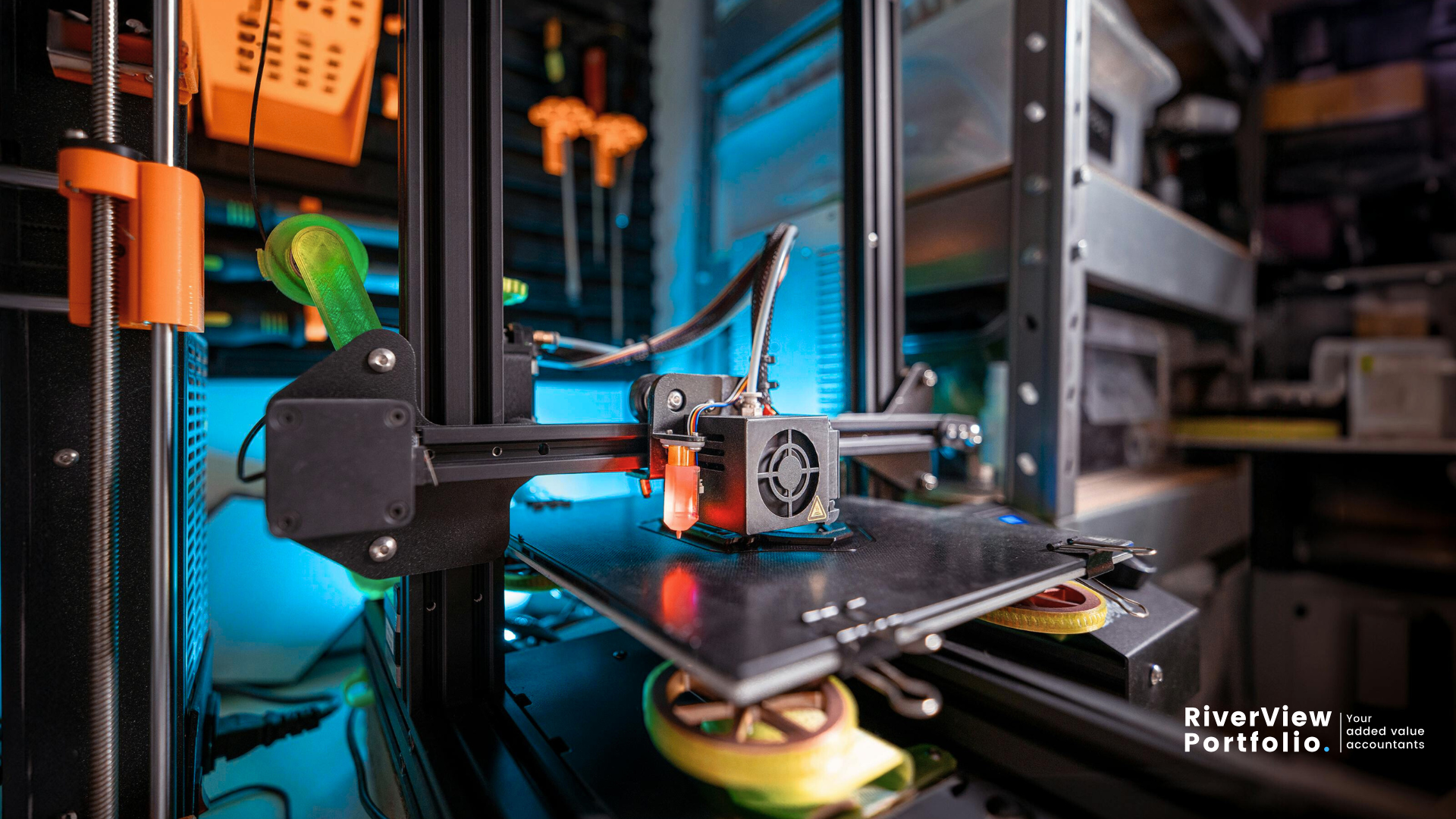 A close-up, dynamic shot of a 3D printer in operation, with its print head actively extruding material onto the build plate. The machine's internal mechanisms, including motors and belts, are visible, illuminated by a blue glow from within. The background is blurred, showing a workshop or lab environment with various tools and equipment, suggesting an innovative and creative space. In the bottom right corner, the "RiverView Portfolio | Your added value accountants" logo is present, indicating the image's relevance to research and development activities and potential R&D tax credit claims. Property Tax Advice Expat Tax Advice PTA ETA Riverview Portfolio