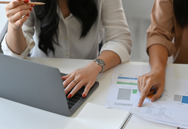 A close-up photograph of two women seated at a desk, looking over documents. The woman on the left is wearing a white shirt and is typing on a grey laptop while holding a pencil in her right hand. She is wearing a wristwatch. The woman on the right is wearing a beige top and is pointing at a paper document with charts and figures, which is next to an open notebook. Property Tax Advice Expat Tax Advice PTA ETA Riverview Portfolio