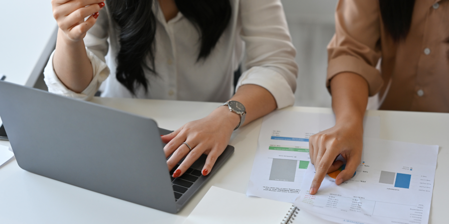 A close-up photograph of two women seated at a desk, looking over documents. The woman on the left is wearing a white shirt and is typing on a grey laptop while holding a pencil in her right hand. She is wearing a wristwatch. The woman on the right is wearing a beige top and is pointing at a paper document with charts and figures, which is next to an open notebook. Property Tax Advice Expat Tax Advice PTA ETA Riverview Portfolio