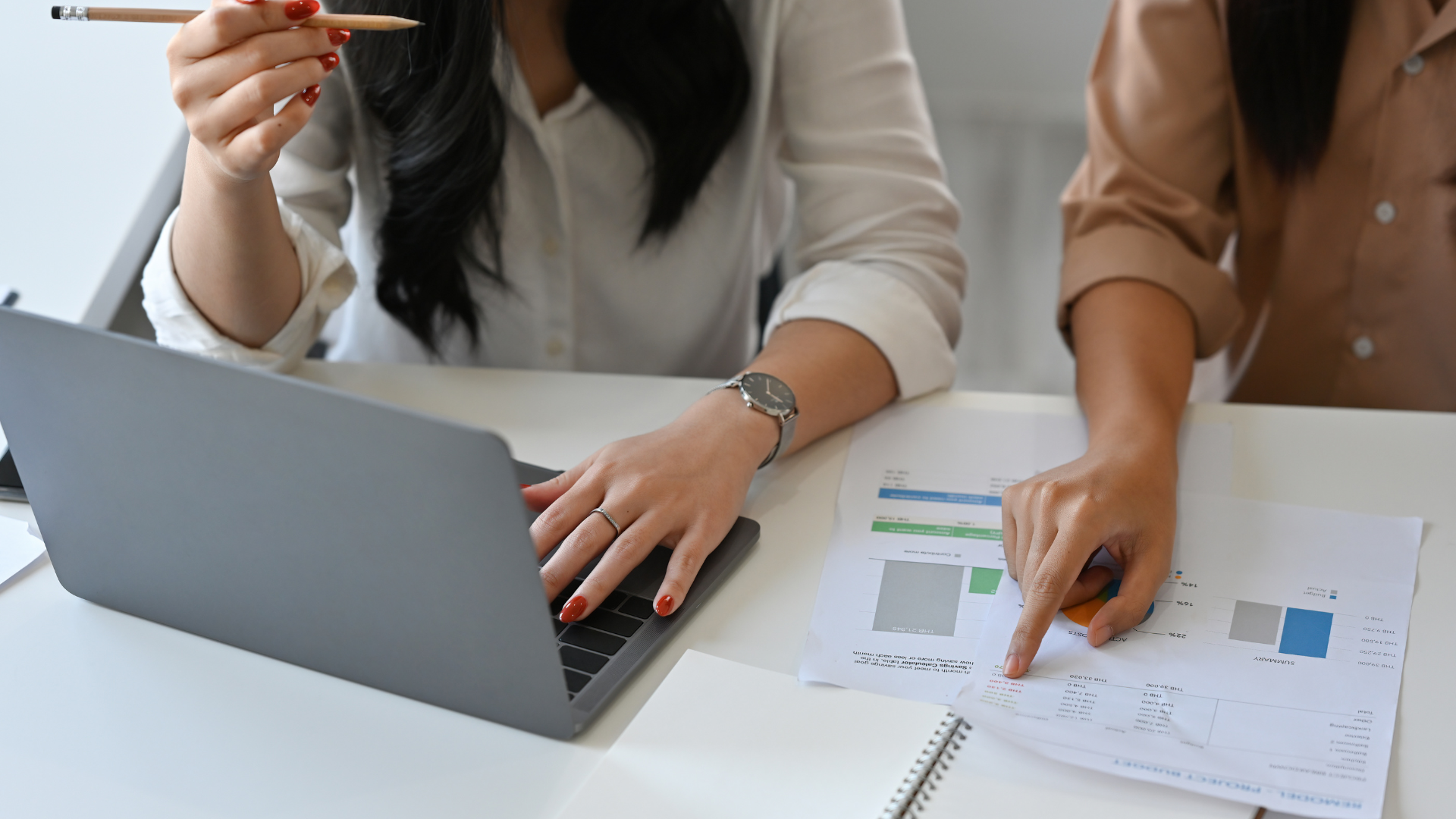 A close-up photograph of two women seated at a desk, looking over documents. The woman on the left is wearing a white shirt and is typing on a grey laptop while holding a pencil in her right hand. She is wearing a wristwatch. The woman on the right is wearing a beige top and is pointing at a paper document with charts and figures, which is next to an open notebook. Property Tax Advice Expat Tax Advice PTA ETA Riverview Portfolio