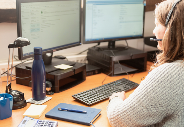 A photograph showing a woman with brown hair, wearing a grey knitted sweater and a headset with a microphone, sitting at a wooden desk and typing on a keyboard. She is looking at two computer monitors displaying what appears to be an email or software interface. On the desk, there is a blue water bottle, a small black desk lamp, a cup with pens and a ruler, a blue notebook, a pen resting on the notebook, and a calculator. Property Tax Advice Expat Tax Advice PTA ETA Riverview Portfolio