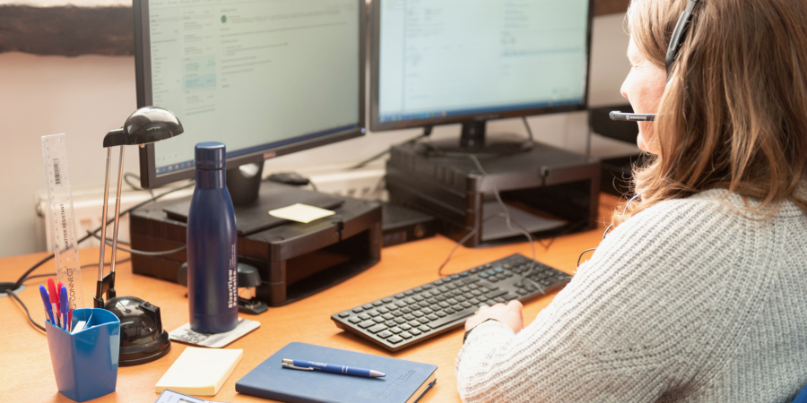A photograph showing a woman with brown hair, wearing a grey knitted sweater and a headset with a microphone, sitting at a wooden desk and typing on a keyboard. She is looking at two computer monitors displaying what appears to be an email or software interface. On the desk, there is a blue water bottle, a small black desk lamp, a cup with pens and a ruler, a blue notebook, a pen resting on the notebook, and a calculator. Property Tax Advice Expat Tax Advice PTA ETA Riverview Portfolio