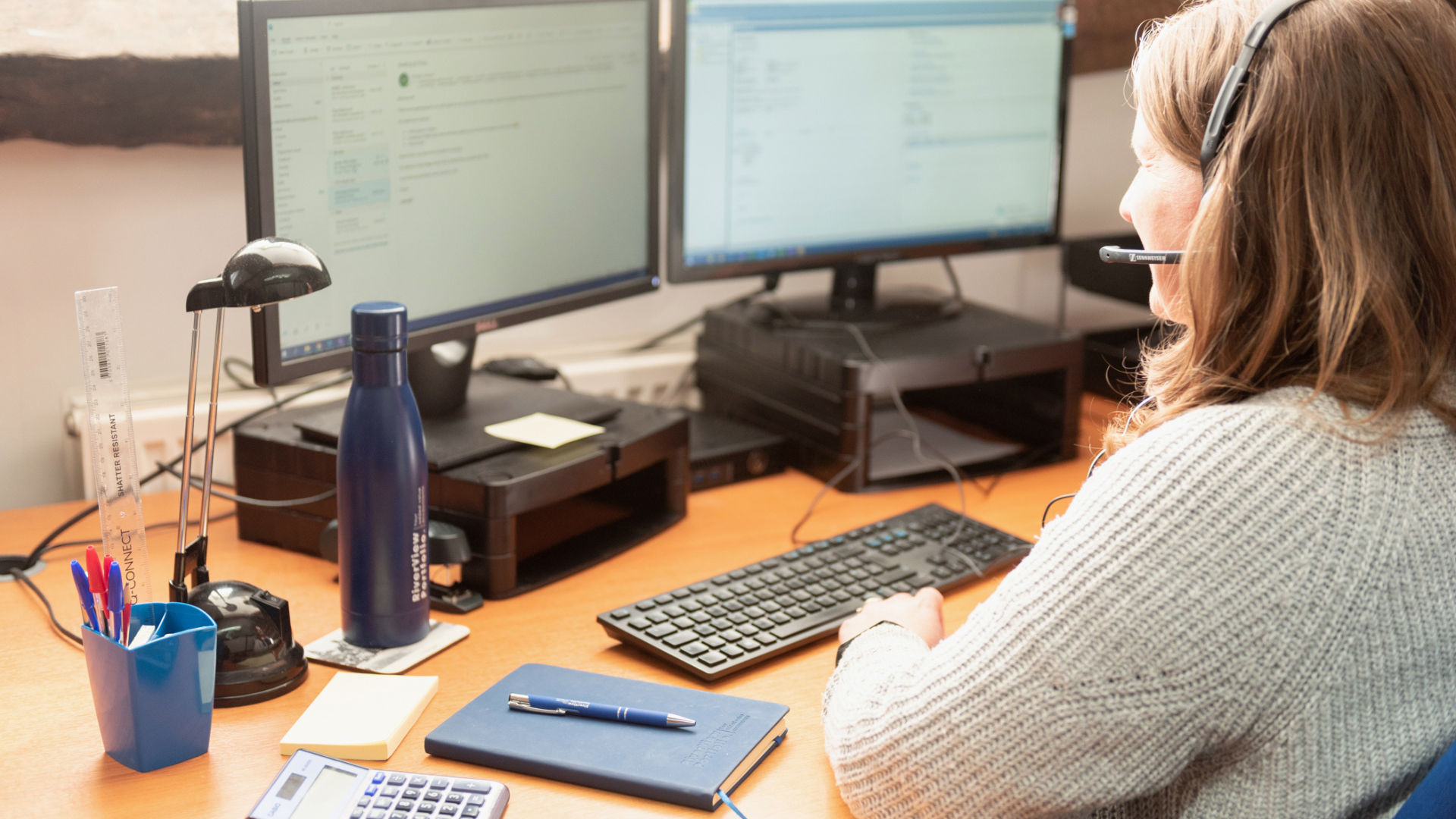 A photograph showing a woman with brown hair, wearing a grey knitted sweater and a headset with a microphone, sitting at a wooden desk and typing on a keyboard. She is looking at two computer monitors displaying what appears to be an email or software interface. On the desk, there is a blue water bottle, a small black desk lamp, a cup with pens and a ruler, a blue notebook, a pen resting on the notebook, and a calculator. Property Tax Advice Expat Tax Advice PTA ETA Riverview Portfolio