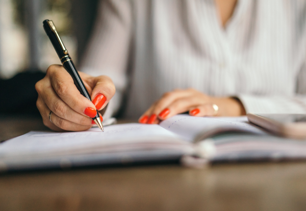 Close-up of a person's hands with red-painted fingernails, holding a black and gold pen and writing in a notebook. The person is wearing a white collared shirt, and a gold ring is visible on their left ring finger. The notebook is open on a wooden table, and a blurred smartphone can be seen to the right. This image illustrates the detailed work involved in company secretarial duties, such as those performed at Riverview Portfolio, where precision and organization are key.
