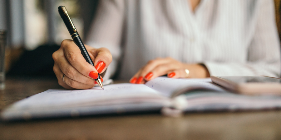 Close-up of a person's hands with red-painted fingernails, holding a black and gold pen and writing in a notebook. The person is wearing a white collared shirt, and a gold ring is visible on their left ring finger. The notebook is open on a wooden table, and a blurred smartphone can be seen to the right. This image illustrates the detailed work involved in company secretarial duties, such as those performed at Riverview Portfolio, where precision and organization are key.
