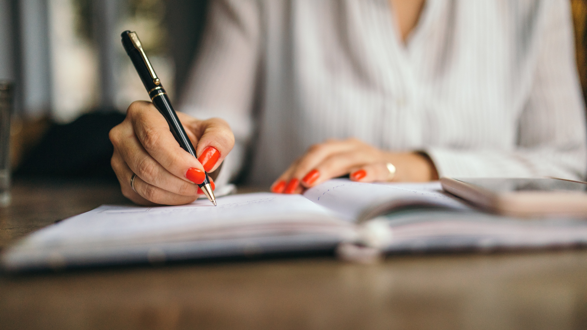 Close-up of a person's hands with red-painted fingernails, holding a black and gold pen and writing in a notebook. The person is wearing a white collared shirt, and a gold ring is visible on their left ring finger. The notebook is open on a wooden table, and a blurred smartphone can be seen to the right. This image illustrates the detailed work involved in company secretarial duties, such as those performed at Riverview Portfolio, where precision and organization are key.