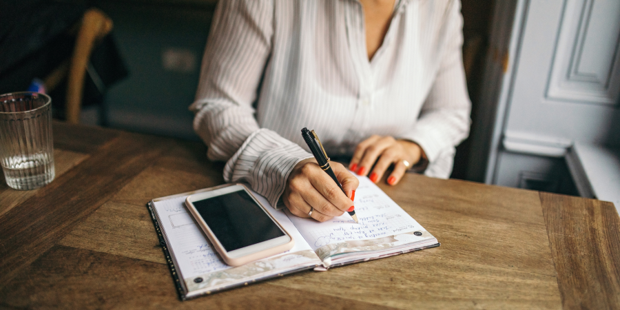 A close-up photograph showing a woman in a white striped blouse writing in an open notebook on a wooden table with a black and gold pen. Her fingernails are painted red. A smartphone with a pale pink case is resting on the left page of the notebook. To the far left, a glass of water is partially visible. Property Tax Advice Expat Tax Advice PTA ETA Riverview Portfolio