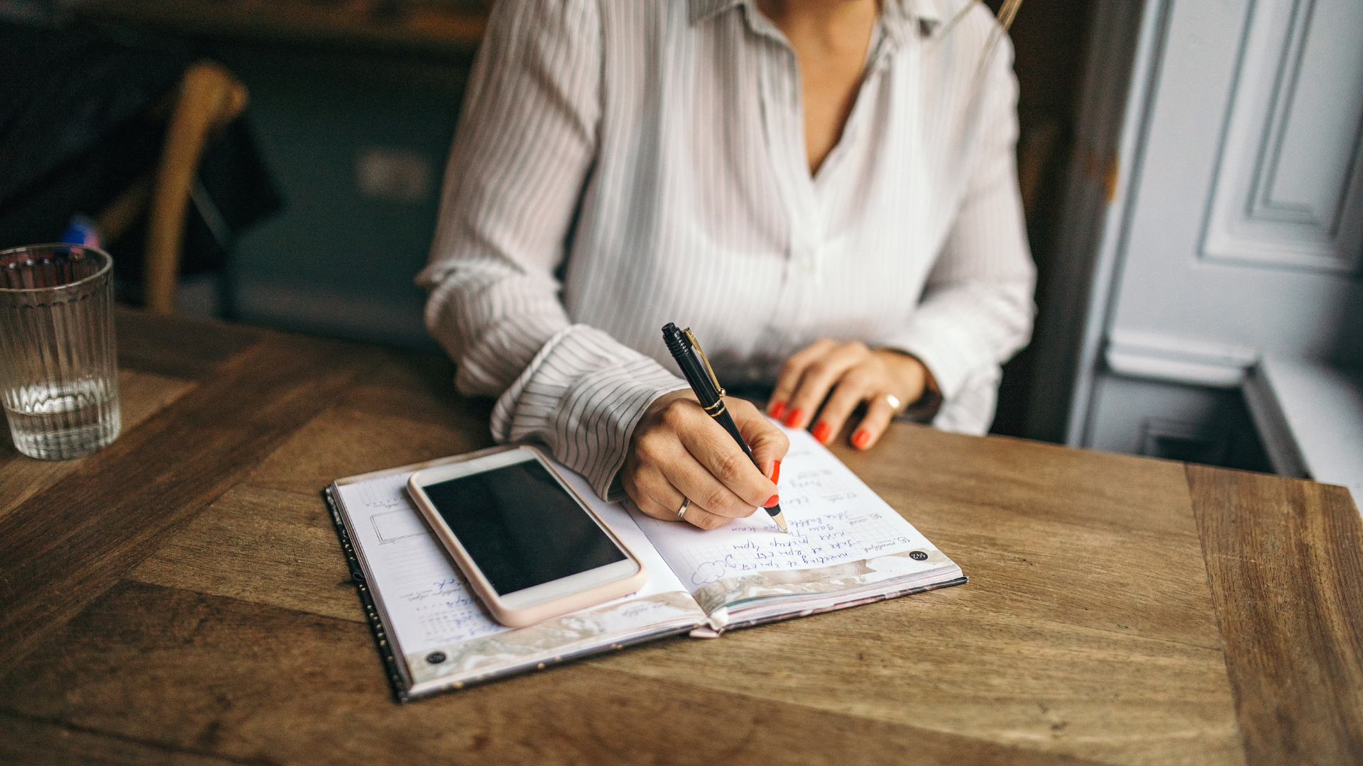 A close-up photograph showing a woman in a white striped blouse writing in an open notebook on a wooden table with a black and gold pen. Her fingernails are painted red. A smartphone with a pale pink case is resting on the left page of the notebook. To the far left, a glass of water is partially visible. Property Tax Advice Expat Tax Advice PTA ETA Riverview Portfolio