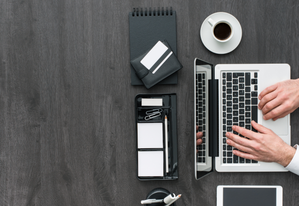 A top-down photograph of a person in a white shirt working on a silver and black laptop on a dark, wood-grain desk. To the left of the laptop are several black and white office supplies, including a black spiral notebook, a white wallet, a desk organizer with a pencil, paper clips, and sticky notes, and a partially visible tablet or phone at the bottom. A white cup of coffee is near the top right. Property Tax Advice Expat Tax Advice PTA ETA Riverview Portfolio
