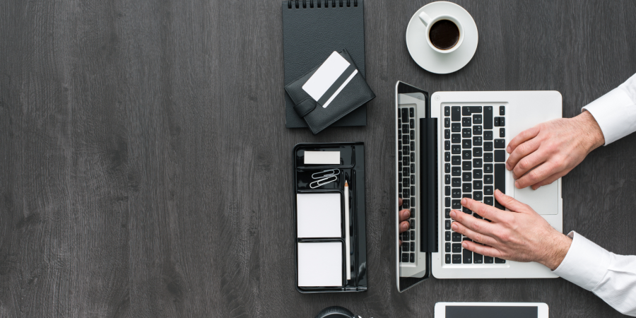 A top-down photograph of a person in a white shirt working on a silver and black laptop on a dark, wood-grain desk. To the left of the laptop are several black and white office supplies, including a black spiral notebook, a white wallet, a desk organizer with a pencil, paper clips, and sticky notes, and a partially visible tablet or phone at the bottom. A white cup of coffee is near the top right. Property Tax Advice Expat Tax Advice PTA ETA Riverview Portfolio