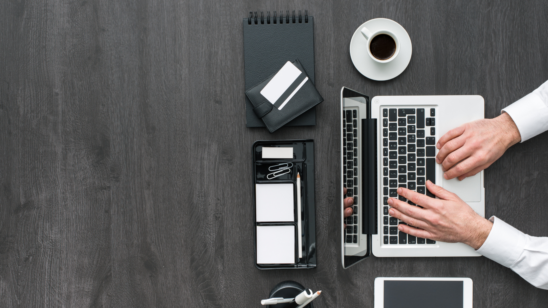 A top-down photograph of a person in a white shirt working on a silver and black laptop on a dark, wood-grain desk. To the left of the laptop are several black and white office supplies, including a black spiral notebook, a white wallet, a desk organizer with a pencil, paper clips, and sticky notes, and a partially visible tablet or phone at the bottom. A white cup of coffee is near the top right. Property Tax Advice Expat Tax Advice PTA ETA Riverview Portfolio